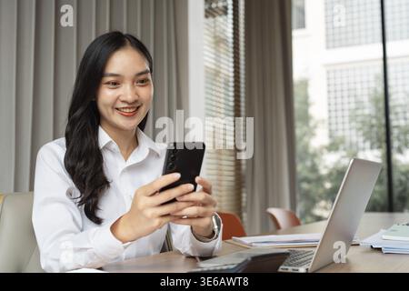 A happy professional woman engages with her smartphone at a modern office desk, surrounded by documents and a laptop, highlighting her multitasking ab Stock Photo