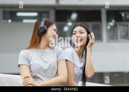 Two joyful students wearing headphones sit together, sharing music and laughter on campus, highlighting their friendship and connection Stock Photo
