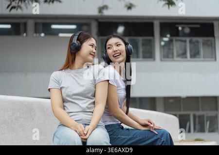 Two diverse university students sitting close together outdoors, smiling and sharing music through headphones, enjoying a moment of friendship on camp Stock Photo
