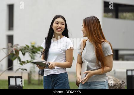 Two diverse university students are walking together on campus, laughing and enjoying their time while carrying books and a laptop Stock Photo