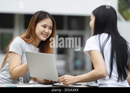 Two diverse university students share a smile while collaborating on a project using a laptop outdoors on campus Stock Photo