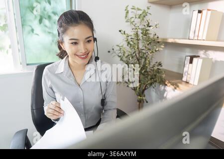 A cheerful woman in a headset interacts with clients while managing documents in her home office, exemplifying remote work success Stock Photo