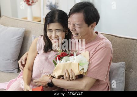 A happy senior couple enjoying Valentine's Day, sharing a joyful moment with flowers on a comfortable sofa Stock Photo