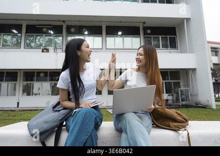 Two diverse female students celebrating a successful moment with a high-five while sitting on a campus bench, sharing joy and camaraderie Stock Photo