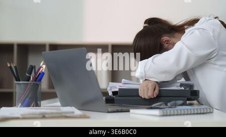 A young woman in a white shirt rests her head on a pile of paperwork at her desk, reflecting the pressures of modern work life Stock Photo