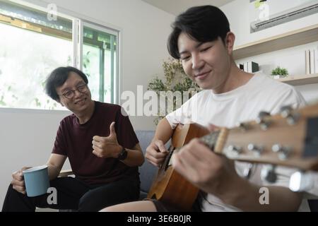 A father smiles and gives a thumbs up while enjoying coffee as his son plays guitar, highlighting their close bond Stock Photo