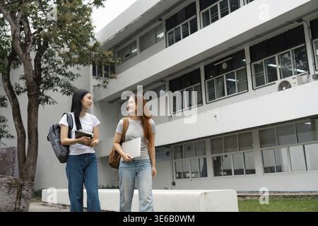 Two university students stroll happily across campus, with one carrying a laptop and the other a tablet, engaging in cheerful conversation Stock Photo