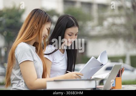 Two diverse university students deeply focused on their studies outdoors, reviewing documents and collaborating next to a laptop Stock Photo