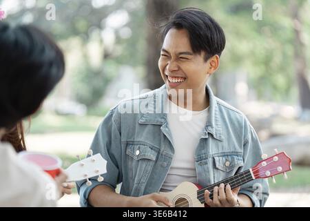A diverse young man joyfully playing the ukulele outdoors with friends in a park, creating a cheerful atmosphere of camaraderie Stock Photo