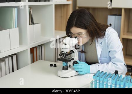 Engaged female scientist in a lab coat and safety glasses, closely examining samples under a microscope in a modern research laboratory Stock Photo