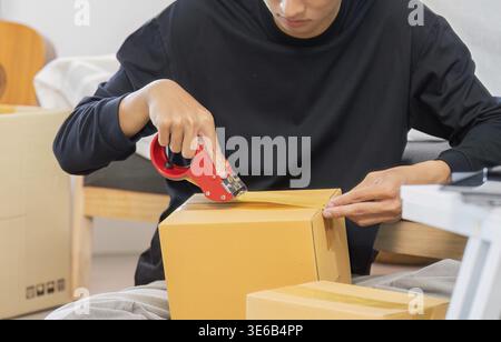 A young man carefully opens a cardboard box using a tape cutter, preparing to unpack items in a modern living space Stock Photo