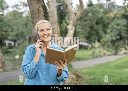 A cheerful senior woman, dressed in a blue shirt, engaging in a phone conversation while reading notes in her notebook near a serene park area Stock Photo