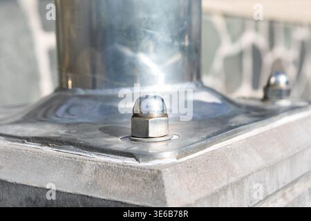 Polished metal pole base secured with bolts and dome caps on a stone surface. Smooth surfaces contrast with the rough stone wall in the background Stock Photo