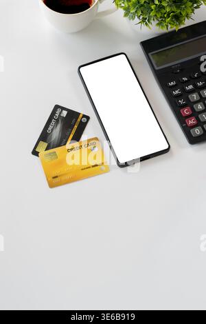 Flat lay of smartphone with blank screen, credit cards, calculator, coffee cup, and plant on white desk, ideal for mobile payment, ecommerce, banking, Stock Photo
