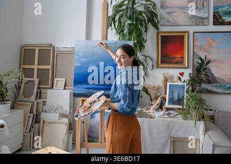 Carefree cheerful woman painter paints picture on easel at studio apartment, art therapy positivity. Stock Photo