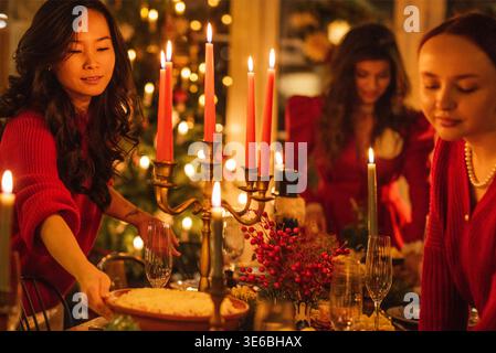 Asian woman in red sweater places dessert on a beautifully set holiday dinner table with candles and decorations, two women are visible in the backgro Stock Photo