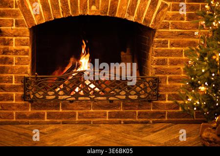 Fireplace with flickering flames set against a brick wall, wooden floor, and a decorated Christmas tree on the right side of the image Stock Photo
