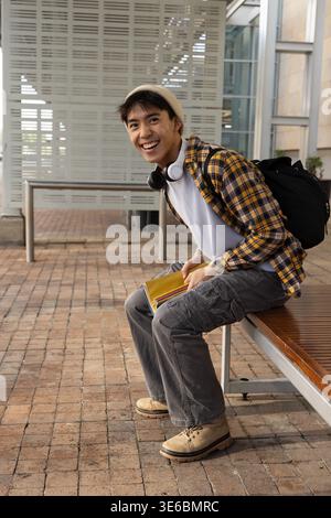 Asian man sitting on wooden metal-framed bench at campus plaza holding yellow notebook smiling Stock Photo