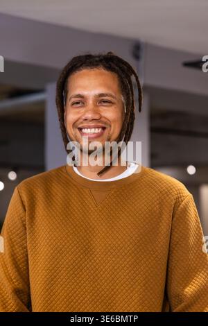 African American man standing, smiling in modern space, wearing mustard pullover over white crew Stock Photo