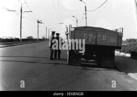 Slaviansk, 1997, on a city bridge. An archive black and white photo capturing a traffic police inspector checking a commercial vehicle. The uniformed officer stands on the asphalt road talking to the driver next to a large dump truck fully loaded with cargo. The truck's wooden tailgate features a hand-painted license plate "92-04 ACN". Trolleybus wires and tall streetlights stretch along the wide, seemingly empty city bridge. A scene of everyday civil service. This is a scene of peaceful life in Donbas Stock Photo
