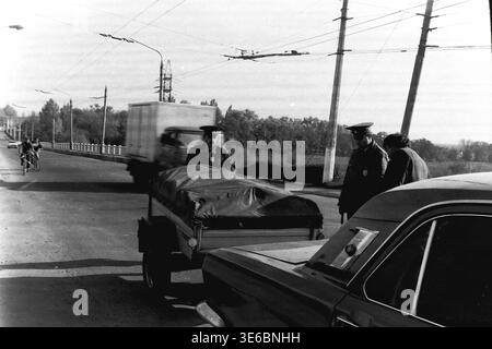 Slaviansk, 1997, on a city bridge road. An archive black and white photo showing traffic police officers conducting a vehicle inspection. Two uniformed inspectors are standing next to a dark passenger car that is pulling a small utility trailer covered tightly with a dark tarp. In the background, a truck drives past them on the wide asphalt road lined with streetlights, overhead trolleybus wires, and autumn trees. A genuine snapshot of everyday road control. This is a scene of peaceful life in Donbas. Stock Photo
