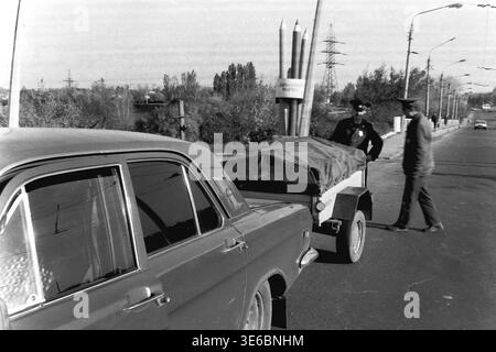 Slaviansk, 1997, on a city bridge road near the pencil factory. An archive black and white photo showing a traffic police vehicle inspection. A male driver is leaning into the open front door of his light-colored passenger car, likely searching for his documents. Two uniformed traffic police officers stand patiently near the small covered utility trailer attached to the vehicle. The famous monument with giant pencils and autumn trees are in the background. This is a scene of peaceful life in Donbas. Stock Photo