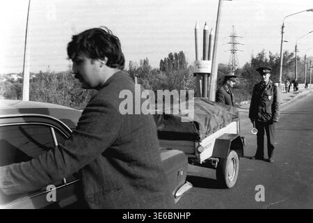 Slaviansk, 1997, on a city bridge road near the pencil factory. An archive black and white photo showing a traffic police vehicle inspection. A male driver is leaning into the open front door of his light-colored passenger car, likely searching for his documents. Two uniformed traffic police officers stand patiently near the small covered utility trailer attached to the vehicle. The famous monument with giant pencils and autumn trees are in the background. This is a scene of peaceful life in Donbas. Stock Photo