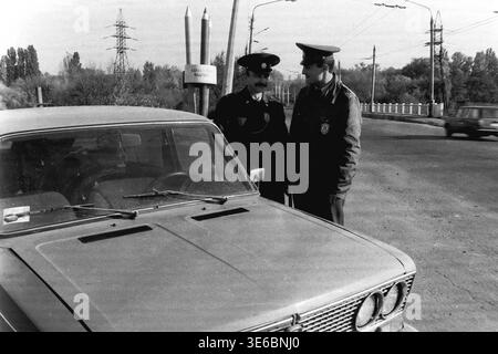 Slaviansk, 1997, on a city bridge near the pencil factory. An archive black and white photo capturing two traffic police officers during a roadside inspection. The uniformed inspectors are standing next to the hood of a light-colored passenger car and talking to each other. The city's famous monument featuring giant pencils, electrical towers, and overhead trolleybus wires are clearly visible in the background against the bright sky. A routine work day. This is a scene of peaceful life in Donbas Stock Photo