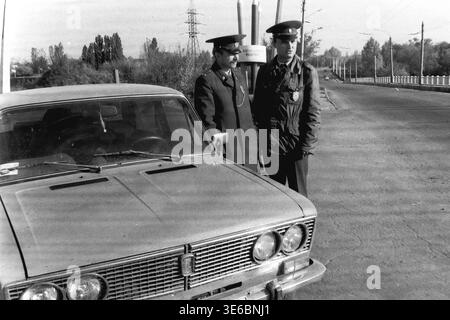 Slaviansk, 1997, on a city bridge near the pencil factory. An archive black and white photo capturing two traffic police officers during a roadside inspection. The uniformed inspectors are standing next to the hood of a light-colored passenger car and talking to each other. The city's famous monument featuring giant pencils, electrical towers, and overhead trolleybus wires are clearly visible in the background against the bright sky. A routine work day. This is a scene of peaceful life in Donbas Stock Photo