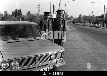 Slaviansk, 1997, on a city bridge near the pencil factory. An archive black and white photo capturing two traffic police officers during a roadside inspection. The uniformed inspectors are standing next to the hood of a light-colored passenger car and talking to each other. The city's famous monument featuring giant pencils, electrical towers, and overhead trolleybus wires are clearly visible in the background against the bright sky. A routine work day. This is a scene of peaceful life in Donbas Stock Photo
