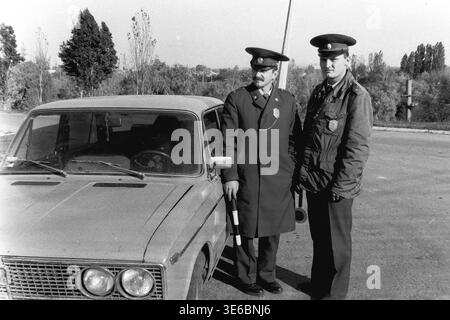 Slaviansk, 1997, on a city bridge road near the pencil factory. An archive black and white photo showing two traffic police officers during a vehicle inspection. The uniformed inspectors are standing next to a light-colored passenger car, holding a striped traffic wand and looking attentively to the right side of the frame. The famous monument with giant pencils, autumn trees, and tall electrical towers are visible in the background. A daily police routine. This is a scene of peaceful life in Donbas. Stock Photo