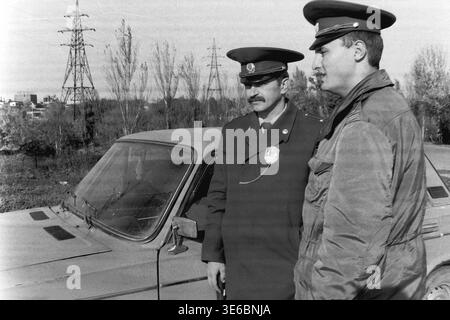 Slaviansk, 1997, on a city bridge road near the pencil factory. An archive black and white photo showing two traffic police officers during a vehicle inspection. The uniformed inspectors are standing next to a light-colored passenger car, holding a striped traffic wand and looking attentively to the right side of the frame. The famous monument with giant pencils, autumn trees, and tall electrical towers are visible in the background. A daily police routine. This is a scene of peaceful life in Donbas. Stock Photo