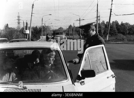 Sloviansk, 1997, city road infrastructure. A uniformed traffic police inspector is carefully checking the driver's documents through the open door of a classic passenger car during a daytime road patrol. In the background, another officer is actively controlling traffic and stopping a heavy truck on the bridge. This authentic archival black and white photograph captures routine law enforcement procedures of the late nineties, everyday vehicle commuting, and the genuine reality of a calm and peaceful Donbas. Stock Photo