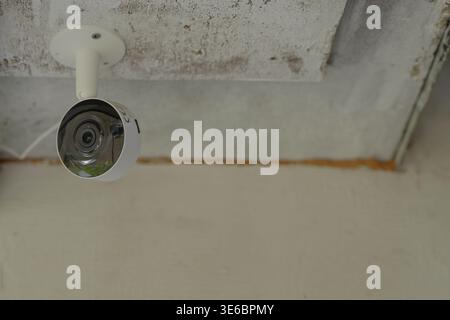 White security camera mounted under a textured ceiling, positioned in the upper corner with visible bracket and reflective lens, set against neutral w Stock Photo