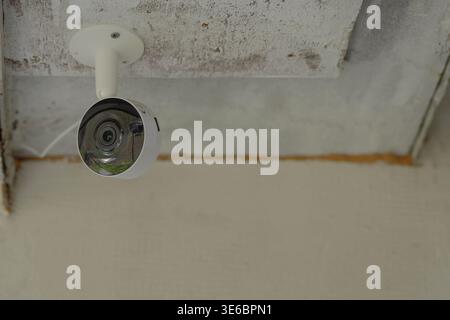 White security camera mounted under a textured ceiling, positioned in the upper corner with visible bracket and reflective lens, set against neutral w Stock Photo