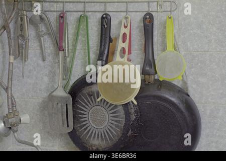 Kitchen corner with hanging utensils and two used frying pans on a metal rack against white tiled wall, showing visible wear, grease marks and everyda Stock Photo