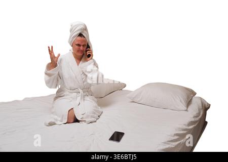 Upset woman in bathrobe talking on phone while sitting on bed. Angry guest complaining about hotel room service. Negative emotion and bad communicatio Stock Photo
