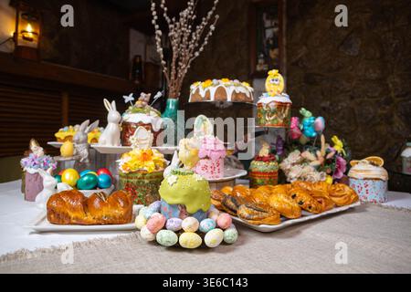 Festive Easter dessert table with colorful cakes, eggs, and floral decorations on stone wall Stock Photo
