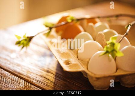 Assorted eggs placed in a carton on a wooden table, featuring a branch with green leaves, highlighting different colors of eggs including brown and bl Stock Photo