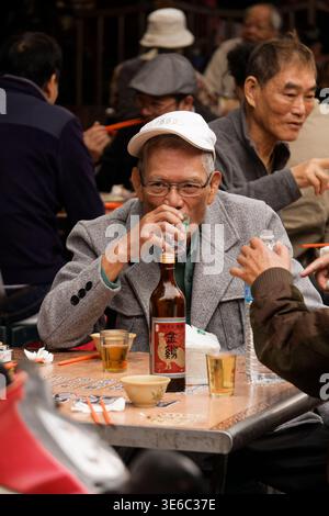 Taipei, Taiwan - March 13, 2024: Elderly man in gray jacket drinks rice wine at outdoor feast near Cisheng Temple, chopsticks and cups on table. Stock Photo