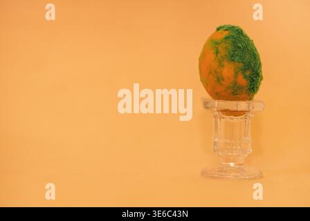 Easter egg featuring a green moss-like texture placed on a glass pedestal, set against a soft orange background, ideal for seasonal decoration Stock Photo