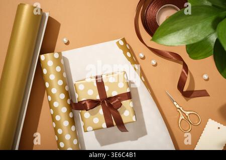 Holiday gift wrapping. Golden box with brown ribbon surrounded by present wrapping items. Stock Photo