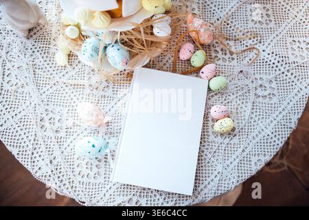 Easter table arrangement featuring a blank notebook and colorful decorative eggs on a lace tablecloth, with a cake stand and festive decor in the back Stock Photo