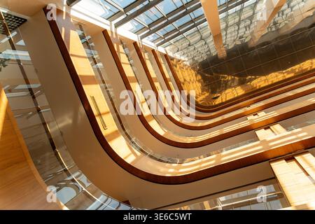Modern atrium interior with curved balconies, glass railings, wooden accents, and a glass ceiling casting sunlight and shadows on reflective surfaces. Stock Photo