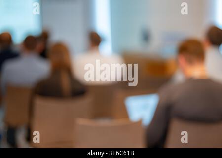 Back view of an audience sitting in chairs at a business conference or seminar, listening to a speaker, with one person using a laptop. Stock Photo