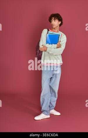 Handsome man with a backpack stands confidently, holding notebooks in front of a colorful wall. Stock Photo
