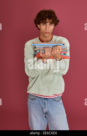 Young man in trendy outfit stands against a pink backdrop, holding colorful notebooks with style. Stock Photo