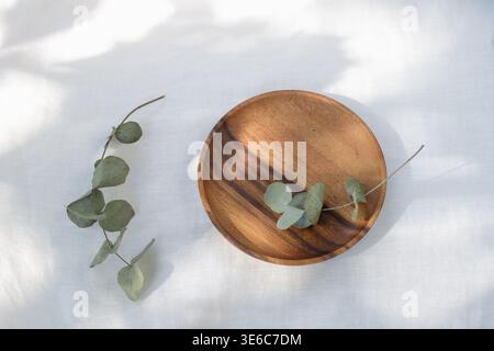 Empty wooden plate with eucalyptus branches on linen tablecloth in sunlight. Summer still life. Mockup floral card. Stock Photo