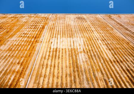 A heavily rusted corrugated metal wall with striking texture, and vertical ridges contracts with the clear blue sky above Stock Photo