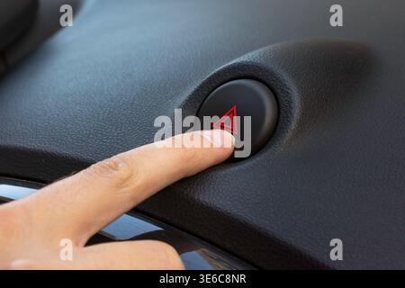 Close-up view of a hand pressing the hazard warning button on a vehicle dashboard, featuring a black interior and a prominent red triangle symbol indi Stock Photo